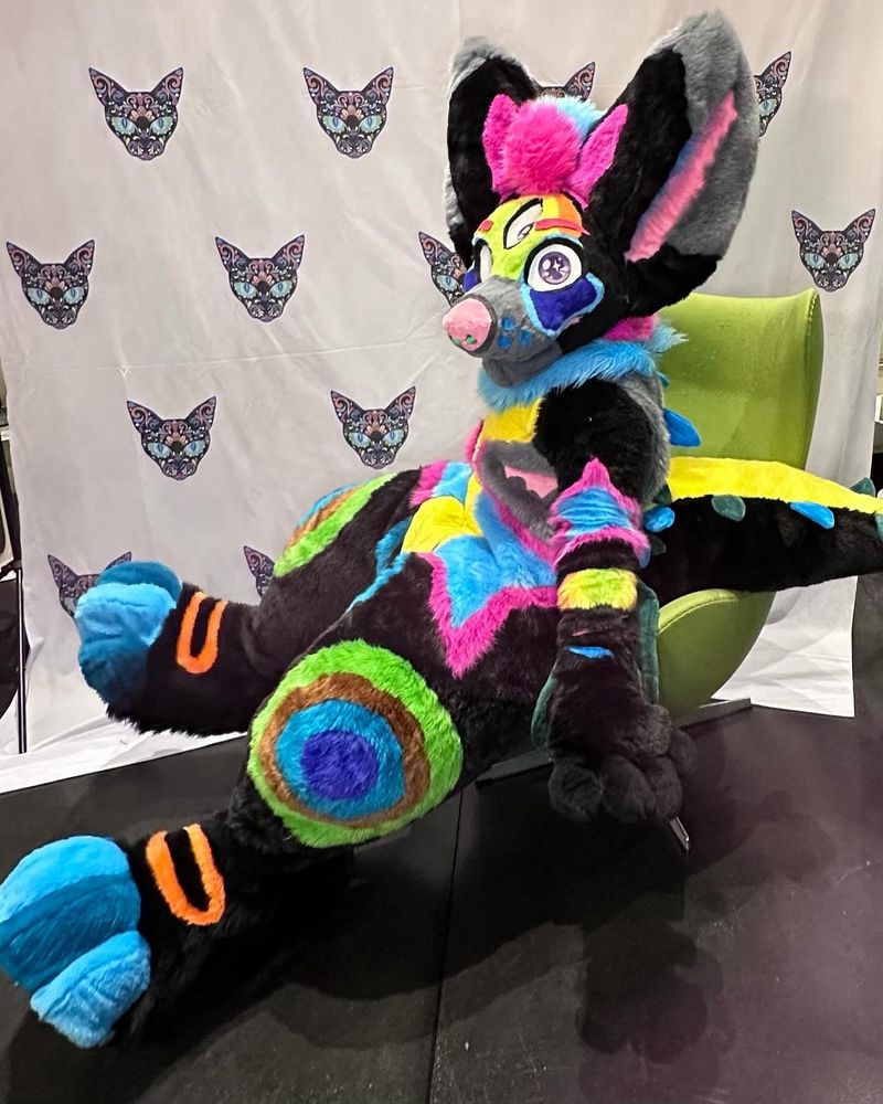 Picture shows Lars, a colorful peacock gatordog fursuit sitting in a green egg chair with a white background patterned with cats.