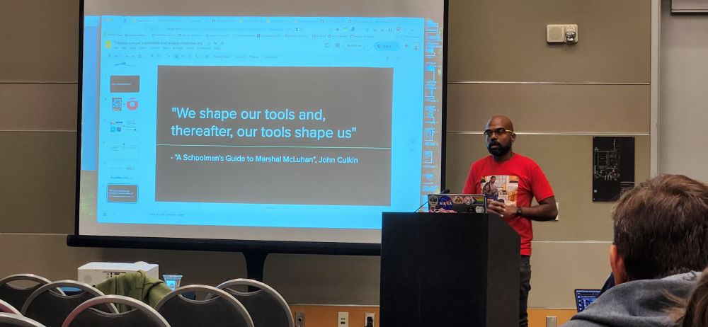A presenter stands at a podium in front of a slide that reads: “We shape our tools and, thereafter, our tools shape us” — A Schoolman’s Guide to Marshall McLuhan, John Culkin.