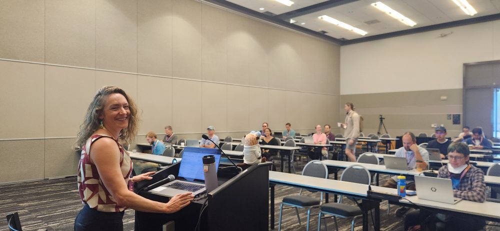 Leah Wasser, Executive Director and Founder of pyOpenSci, smiles at the camera while standing at a podium with a laptop and microphone in front of her. Behind her, participants sit at tables with laptops in a conference room, attending a session at SciPy 2025.