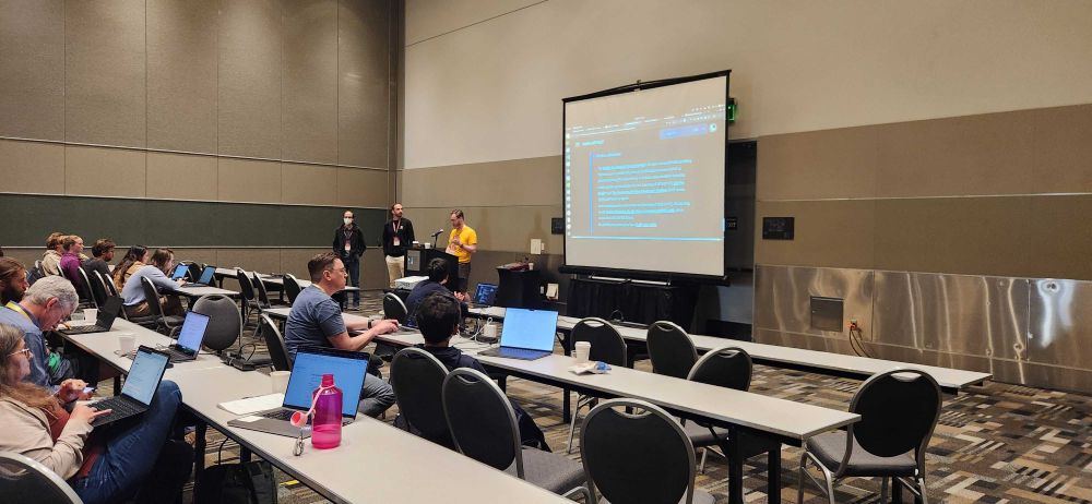 A workshop room with participants seated at rows of tables, working on laptops. Three presenters stand at the front near a projection screen showing a slide about Pixi workflows.