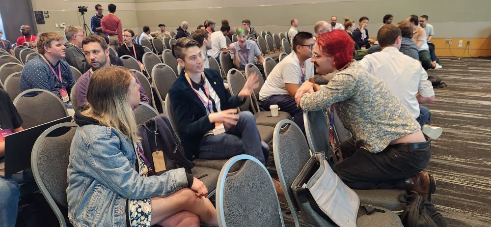 Conference attendees in lively discussion, seated in small groups in a room filled with chairs and people talking and laughing.