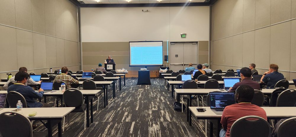 A conference room with attendees seated at tables, working on laptops during a Python packaging workshop at SciPy 2025. A presenter stands at the front podium next to a large screen displaying code examples. The room has a quiet, focused atmosphere with people engaged in hands-on learning.