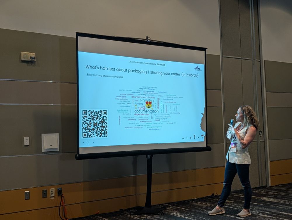Leah Wasser, Executive Director of pyOpenSci, speaks into a microphone while pointing to a word cloud on a slide about challenges in packaging and sharing code. The word cloud highlights “documentation” and “time.”