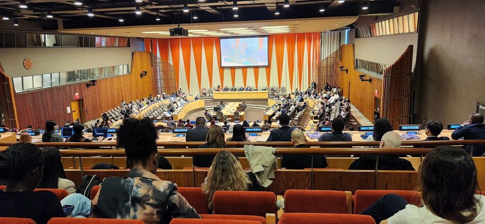 Wide-angle view of a high-level meeting at the United Nations, with delegates seated in a large, wood-paneled conference hall beneath a central screen