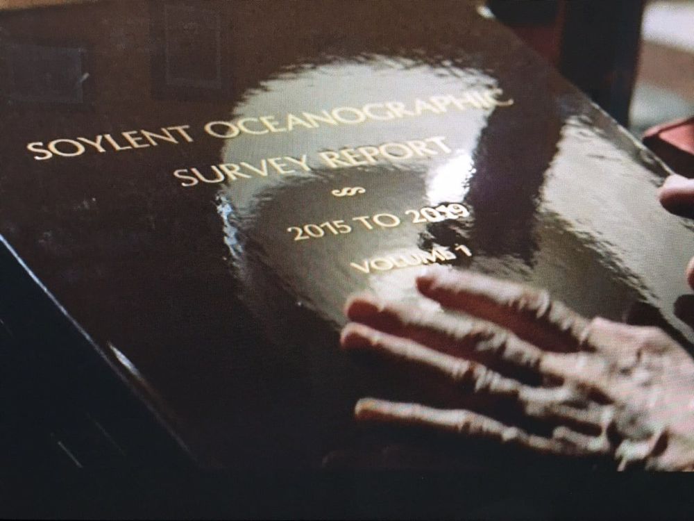 An oversized hardcover Soylent Oceanographic Survey Report 2015-2019 with an aged hand hovering above the cover. It's an expensively bound book with a high gloss cover and it's title is lightly embossed in gold. A screenshot from Soylent Green.