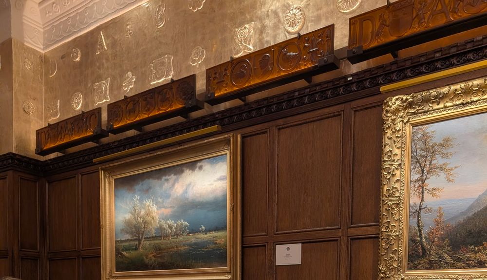 One and a half gold framed pastoral oil paintings hanging on the wood paneled wall of the dining hall beneath a row of tastefully soffited lighting panels that intersect half way down across a vaulted ceiling with gently sweeping plaster coves and brushed gold embossed wall panels.