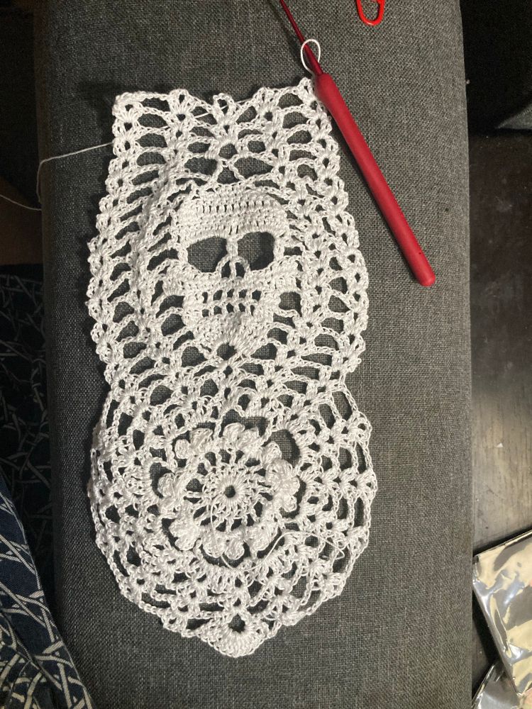 a lacy doily made of white thread with a skull motif. The crochet hook is in the top right corner bc the work is still in progress.