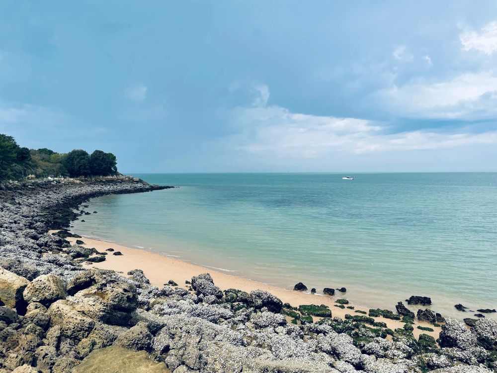 Une plage à l'île d'Aix, au premier plan des rochers puis le sable et la mer aux reflets verdoyants, le ciel est nuageux et la lumière est belle.