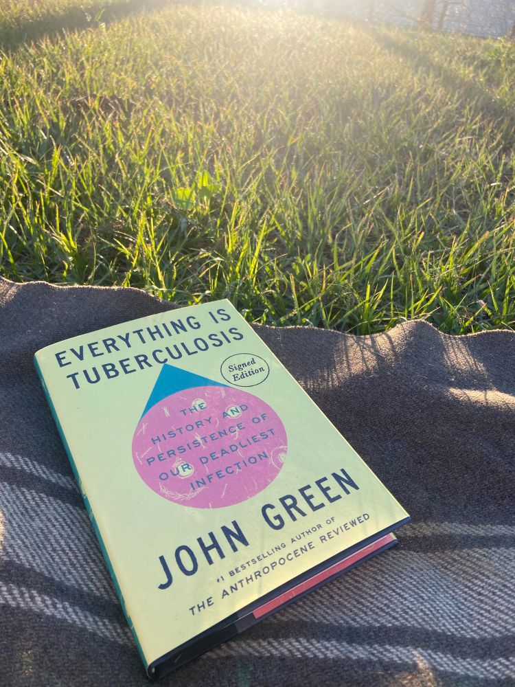 The book Everything is Tuberculosis by John Green sits on a blanket on the grass.