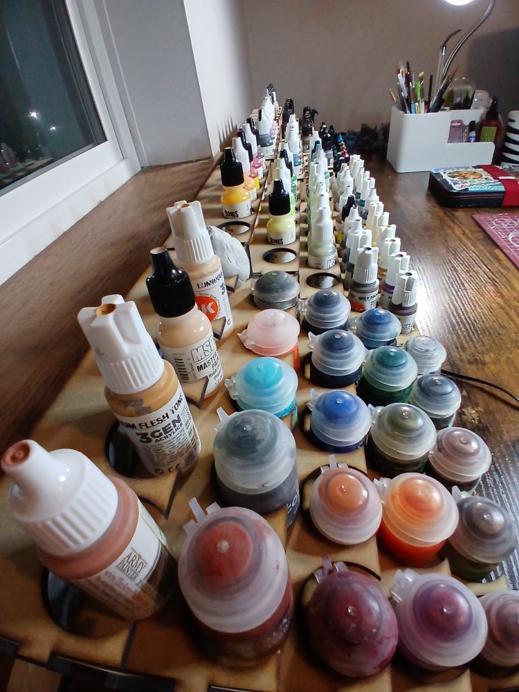 .5 view of the paint rack, to make it appear very long. up close are some flesh tones and a rainbow assortment of paint pots. the rows of paint bottles stretch away. at the far end is a cluttered desk lamp.