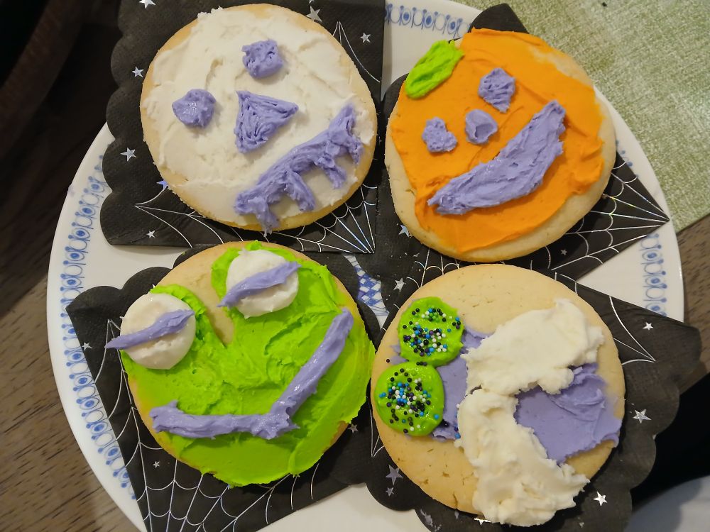 four sugar cookies decorated with big blobs of buttercream icing in white, orange, purple, and green. there is a frog, a fly, a mummy, and a jackolantern