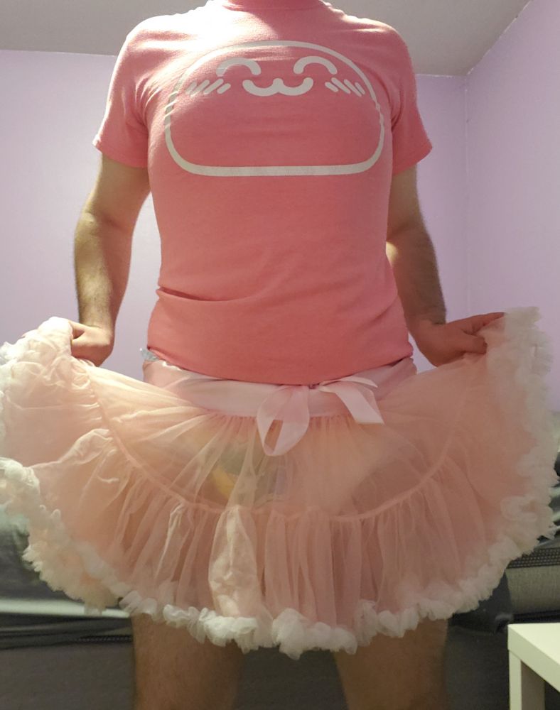 Me dressed in a pink tee with a kawaii blob character printed on it, holding up the sides of a frilly pink & white diaper cover/skirt, which has a pair of Alphagatorz barely hidden underneath