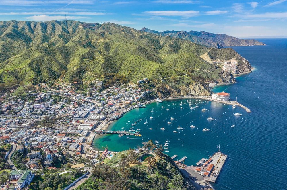 An aerial photograph taken from just above the bluffs that overlook Avalon Bay, showing the city of Avalon (the only incorporated city on the island, the Southernmost within within Los Angeles County). Notably includes Catalina Casino at the Northern end of the boardwalk, a project funded by William Wrigley Jr. (the Chicago gum magnate) in 1928. 