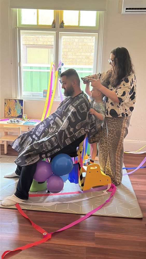 Omar sitting on a chair festooned with balloons and ribbons. His son is standing on a step-stool behind him holding a shaver, while his mum guides his hand. 