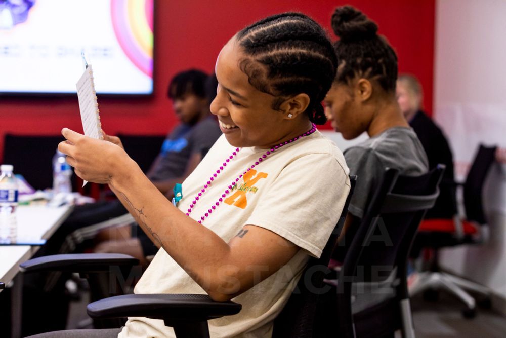 Person smiling, leaning back in a chair, holding a notepad. People are sitting around a table in the background.