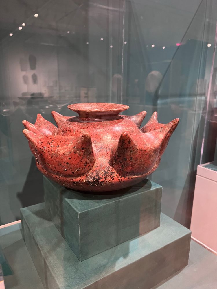 Image of a reddish orange stone sculpture in the shape circular vessel with leaves pointing out from its side.