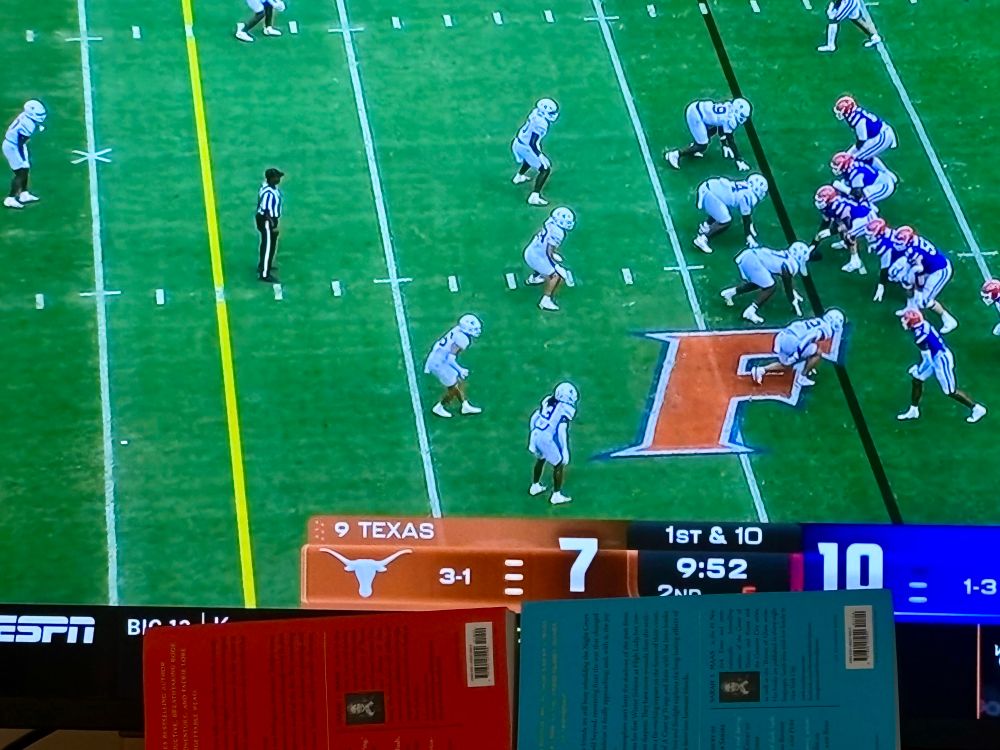 A close shot of a TV playing the Texas-Florida football game. Two paperback books are propped up to cover the bottom of the screen where ESPN shows score updates for other games.