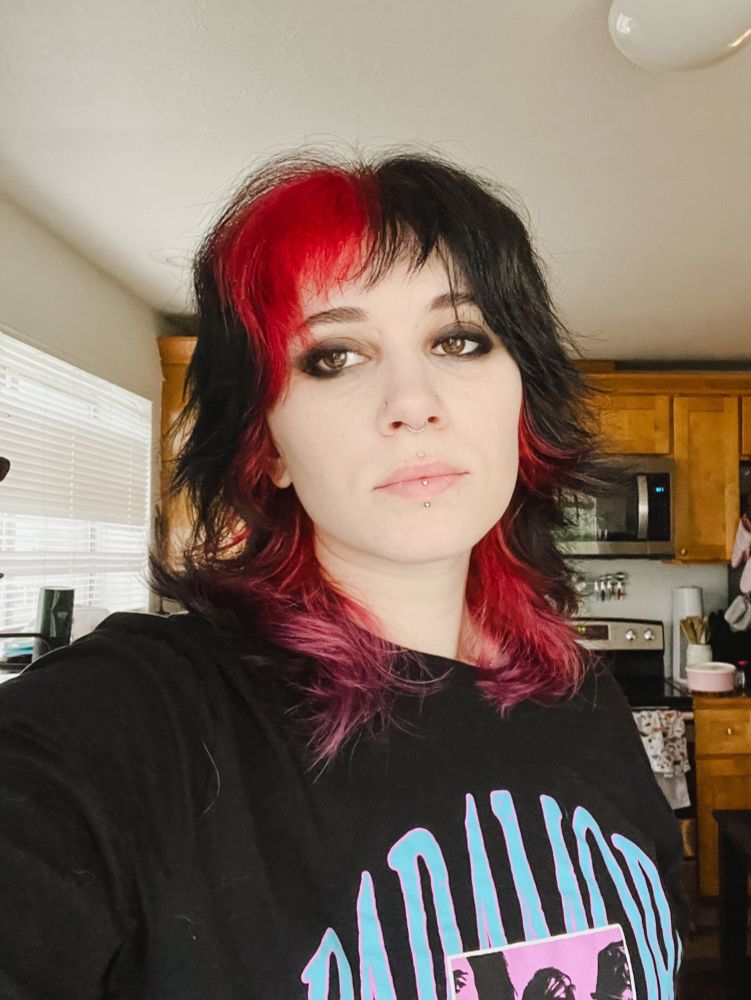 white femme person with black and red hair and lots of eyeliner looking into the camera