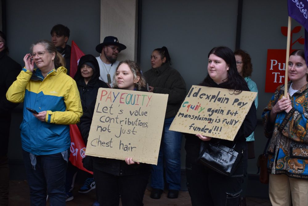 Women holding signs. One reads 'Pay equity. Let's value contributions, not just chest hair' and 'I'd explain pay equity again but mansplaining is fully funded!!!'