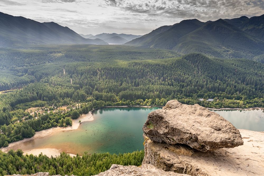 View toward a mountain valley and lake from near the edge of a cliff with a boulder in the foreground. Broken clouds overhead and an expanse of conifers beyond a blue and green lake.