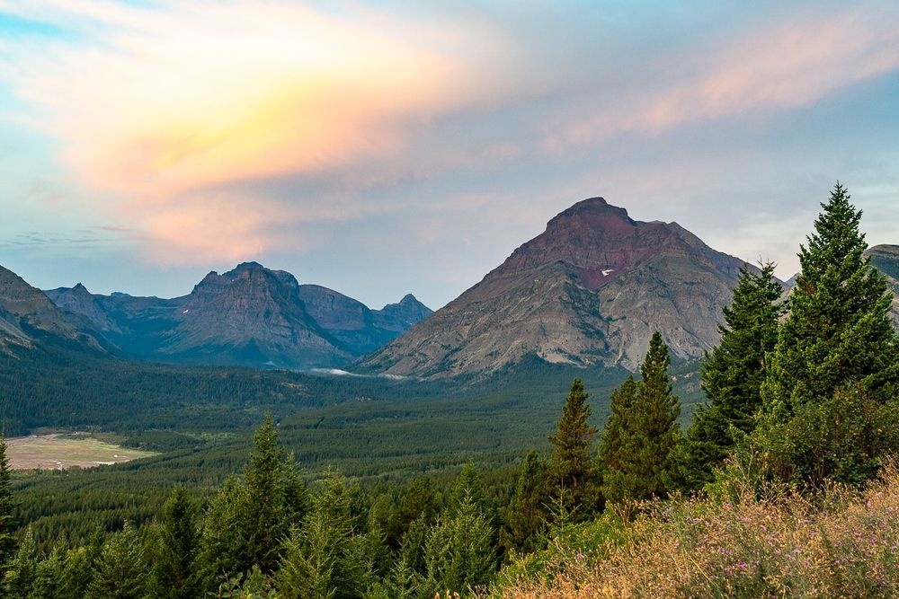 Photo shows two mountains in the background, with the mountain on the right larger and closer, under a sunrise-lit high cloud. In the foreground evergreens line the roadside pullout from where the photo was taken, and stretch into the distance to the base of the mountains.