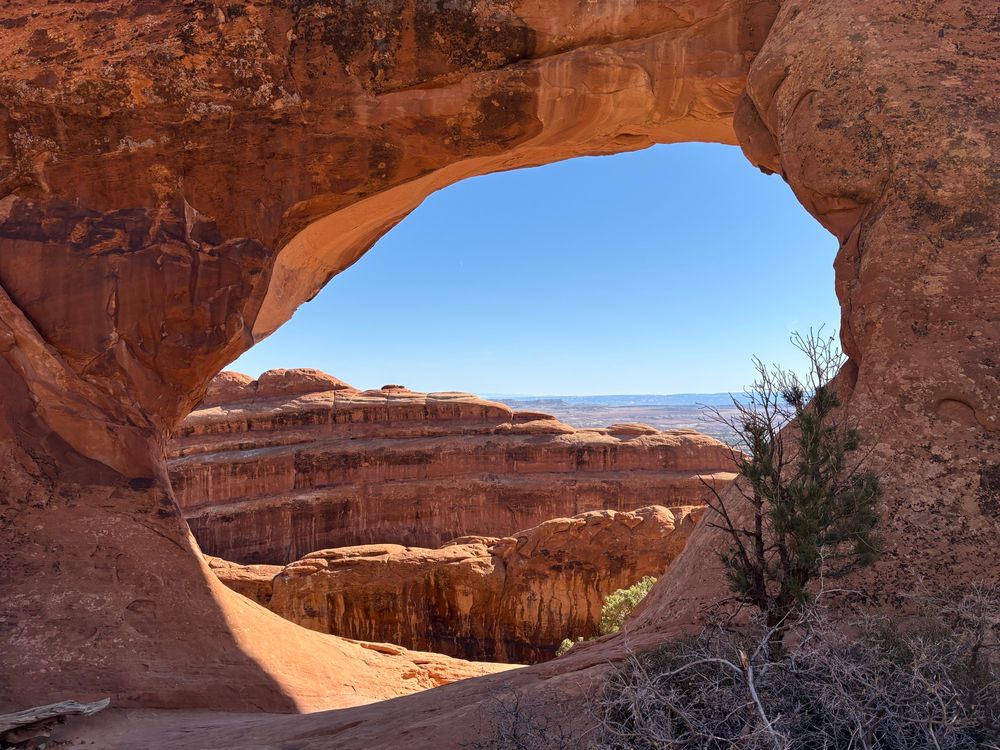 View looking east through a tan and brown natural arch, with rocky fins in the foreground and valley in the distance under a blue sky.