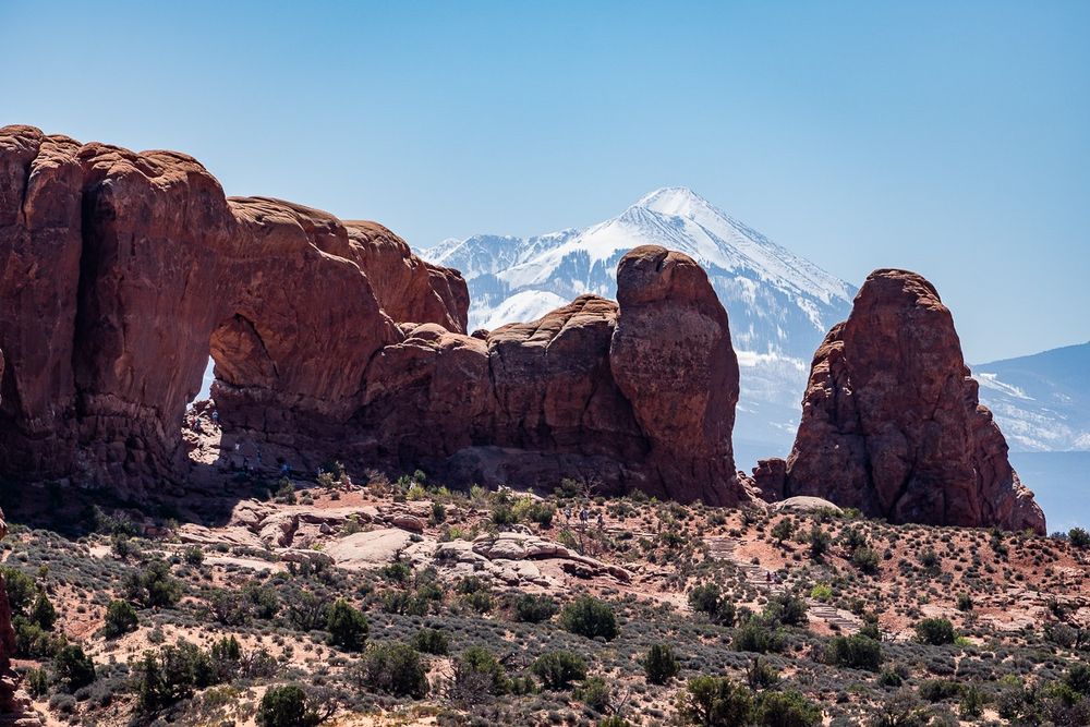 A cone shaped snow capped mountain is seen behind rock formations of Arches National Park. 
