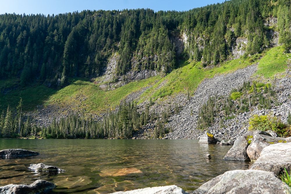 Photo of Upper Granite Lake, with the south shore lined by evergreen topped cliffs with a talus slope base from lake edge to half way up the cliffs. Some trees are growing in the talus at the lake edge and other greenery starts about halfway up the slopes. Several large rocks are in the foreground at the near side lake edge. The distant water is a sea green color, with the near side translucent showing brown rocks just below the surface. A blue sky is overhead at the top of the frame. Morning light gives dark shadows on the trees above the cliffs.