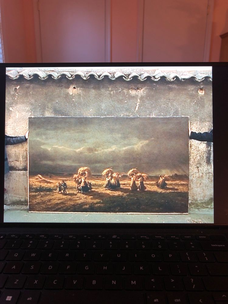 An image of a still from a movie being played on a laptop. The still is a painting resting on a wall. The painting depicts several gleaners in a field hauling wheat and a stormy sky above them. 