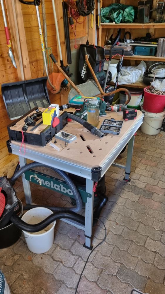 Ein Werktisch in einem Gartenhaus. Die Tischplatte liegt voller Werkzeug und Montagematerial. Mittendrin steht noch eine Trinkflasche.