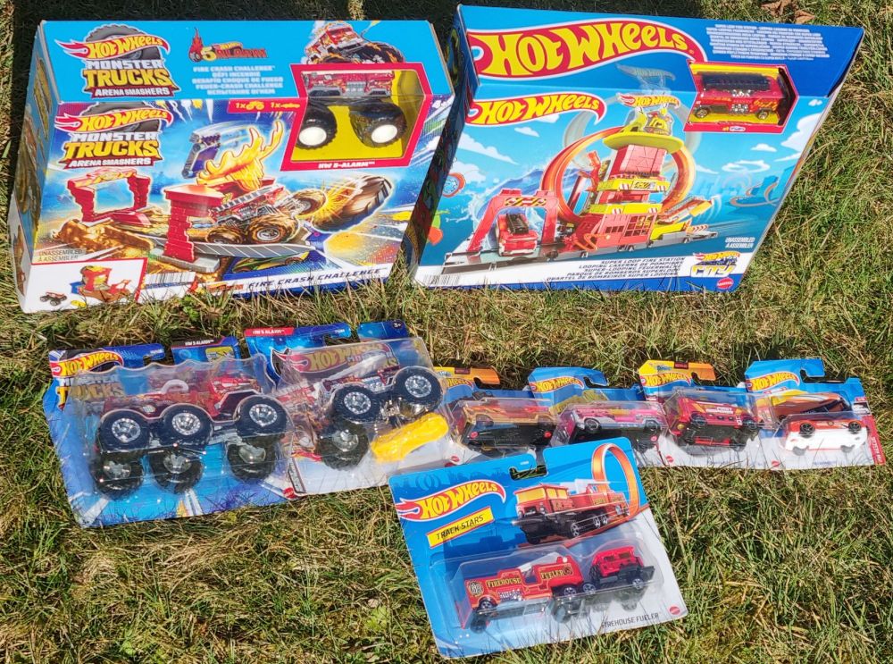 Mehrere originalverpackte Spielzeuge der Marke Hot Wheels auf einer Wiese liegend. Eine Feuerwache mit dem Drehleiterfahrzeug namens „5 Alarm“, ein Feuerstunt-Spielset mit „5 Alarm“ als Monster Truck. Davor liegen 4 weitere Varianten von „5 Alarm“, davon ein zweiachsiger und ein dreiachsiger Monster Truck. Außerdem 3 weitere Feuerwehrfahrzeuge.