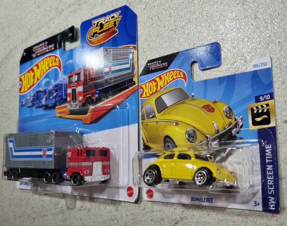 Die besagten Fahrzeuge wie man sie aus der ersten Generation Transformers kennt: Optimus Prime als rote cab-over-engine Sattelzugmaschine mit silbergrauem Auflieger, Bumblebee als gelber VW Käfer. Beide Spielzeuge sind noch originalverpackt.