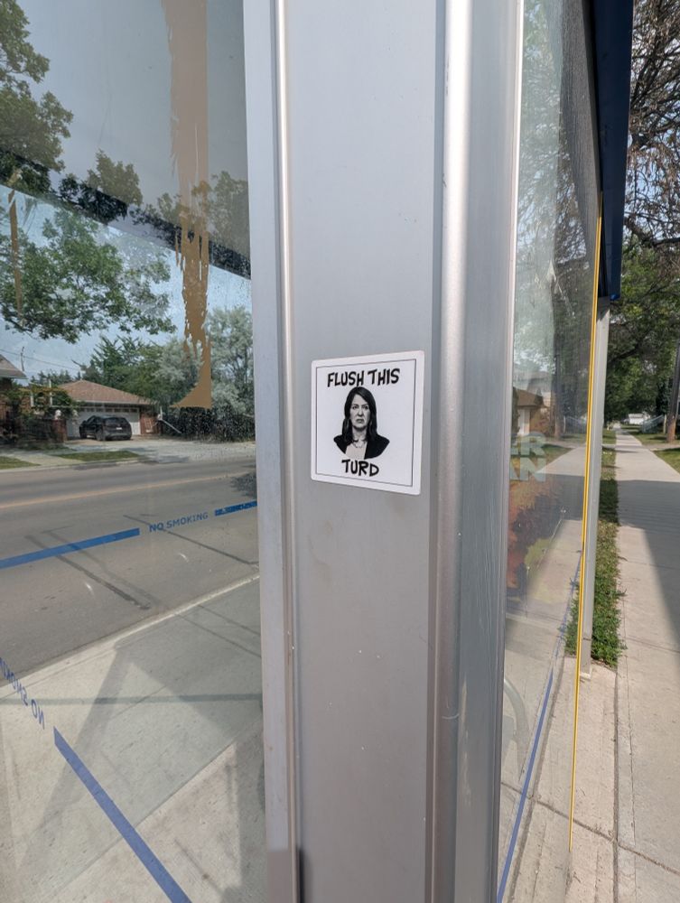 Photo of the black and white sticker on a bus stop. The sticker says "Flush This Turd" around a black and white photo of  piece of shit Alberta Premiere Danielle Smith