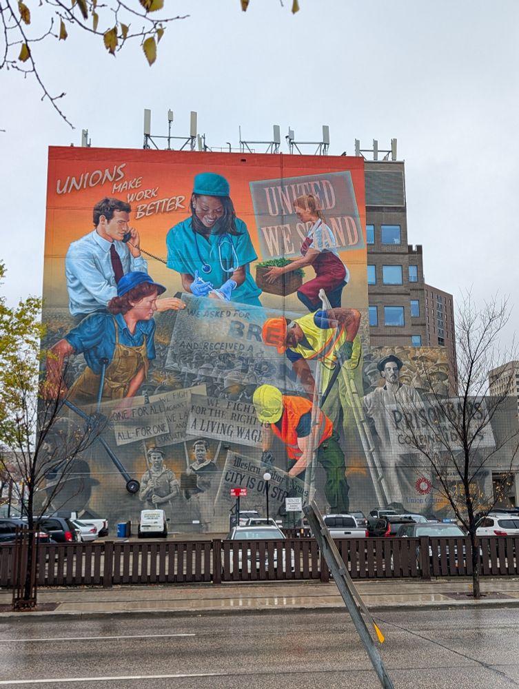 a mural on a large building. The top in bright colour showing people of various genders & ethnicities doing various jobs. It slowly fades out to a grey scale on the bottom which depicts ppl striking & protesting for better working conditioned. Their signs say things like "Prison bars confine ideas"We asked for bread and they gave us a stone," 

At the top it says "Unions make work better" & " United We Stand" 