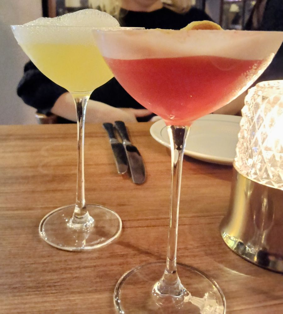 Two longstemmed cocktails next to each other, one lemon yellow and topped with a mound of frothy bubbles, the other rosy pink with foam and a dried citrus slice.