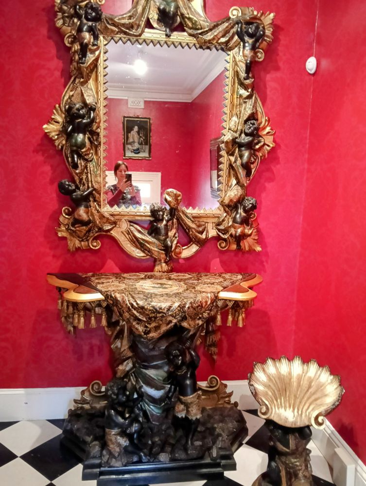 Against a vibrant red wall and on black and white check tile stands a wildly ornate side table and big mirror. Both are cluttered with black cherubs amidst gilded decoration.