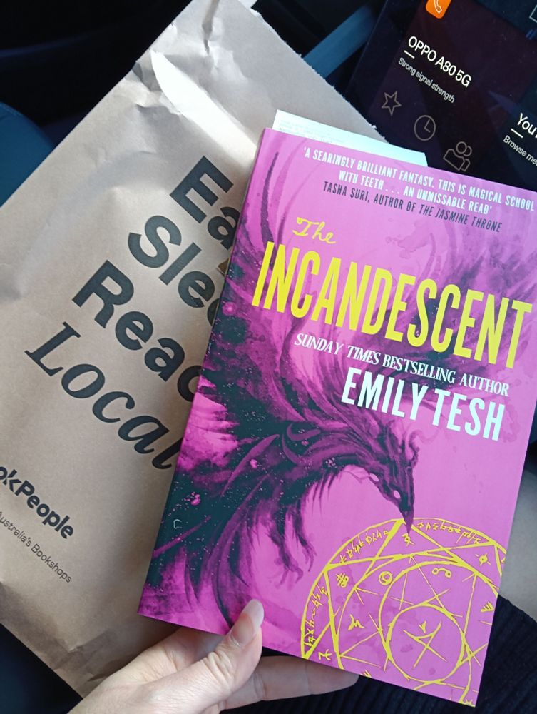 A copy of THE INCANDESCENT by Emily Tesh in all its pink glory atop a paper bag reading "Eat sleep read local".