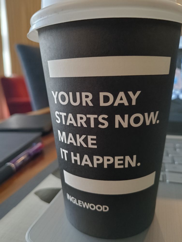Close-up of a disposable coffee cup, black with white text in all caps reading: Your day starts now. Make it happen.