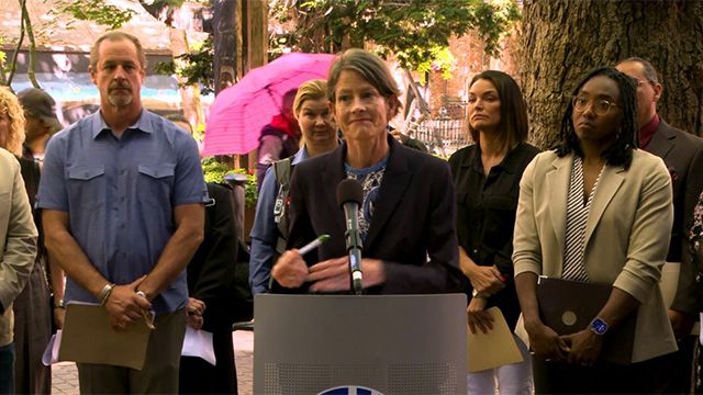 Council President Sara Nelson stands at podium outdoors in black blazer, people gather around and behind