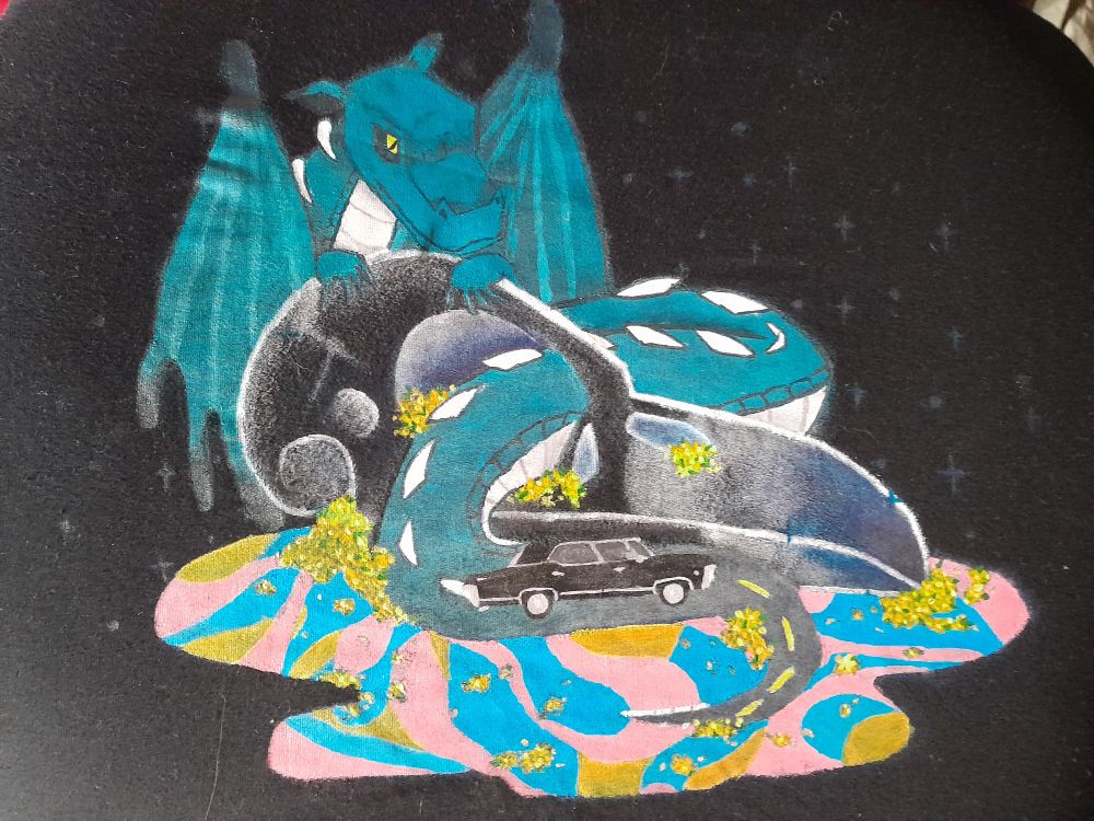 A t shirt on a chair so you the graphic is flat. It is a black shirt with a blue dragon wrapped around a bird skull. The end of the dragons tail fades into a road with a 67 Chevy Impala from the tv show supernatural. 