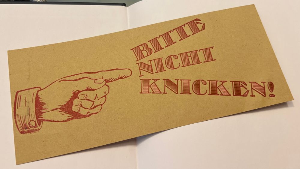 Foto von einem Druck von Guido Kühn:

Auf braunen Umschlagpapier ist eine Hand in dunkelroter Farbe gedruckt, die nach rechts zeigt, wo aufgefächert Aber untereinander steht:

‚BITTE
NICHT
KNICKEN!‘

Das besondere nicht sichtbare daran ist, dass man den Druck mit den Fingern spüren kann!