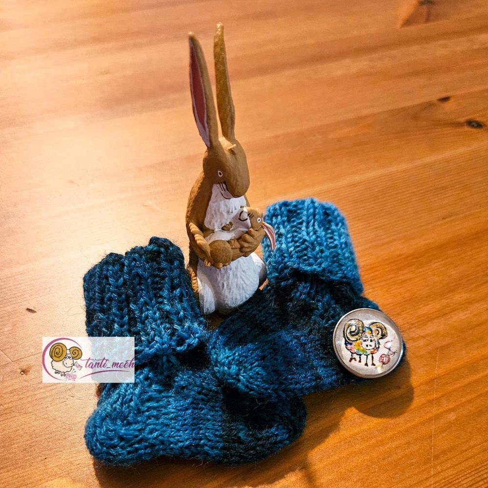 Ganz kleine Socken (ca. 4 cm lang) in Seegrüntönen. Zwischen den  beiden Sockenspitzen steht eine Hasenfigur mit Hasenbahy auf dem Arm. (Toniesfigur "Weisst du eigentlich, wie lieb ich dich hab"). Am rechten Fersenrand liegt ein Druckknopf mit einem bunten, handgemalten Schaf.
