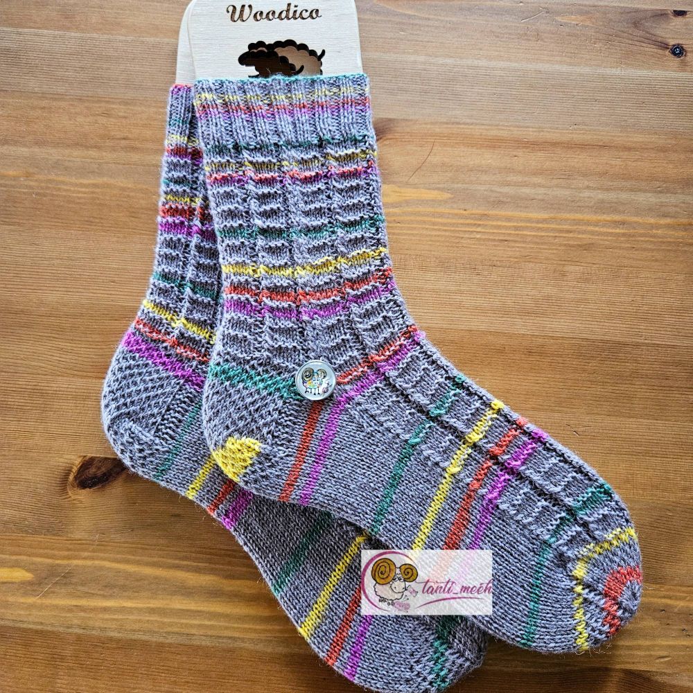 Gestrickte Socken auf Sockenbretter gespannt in beige mit Streifen in grün, orange, pink und gelb. Das Strickbild zeigt am rechten Fussrand eine Gestrickte Kordel. Auf dem Fussrücken sind kleine Rechtecke zu erkennen. Auf Höhe des Fersenanfang liegt ein Druckknopf mit einem bunten, handgemalten Schaf. Die Socken liegen auf einem Kiefertisch.