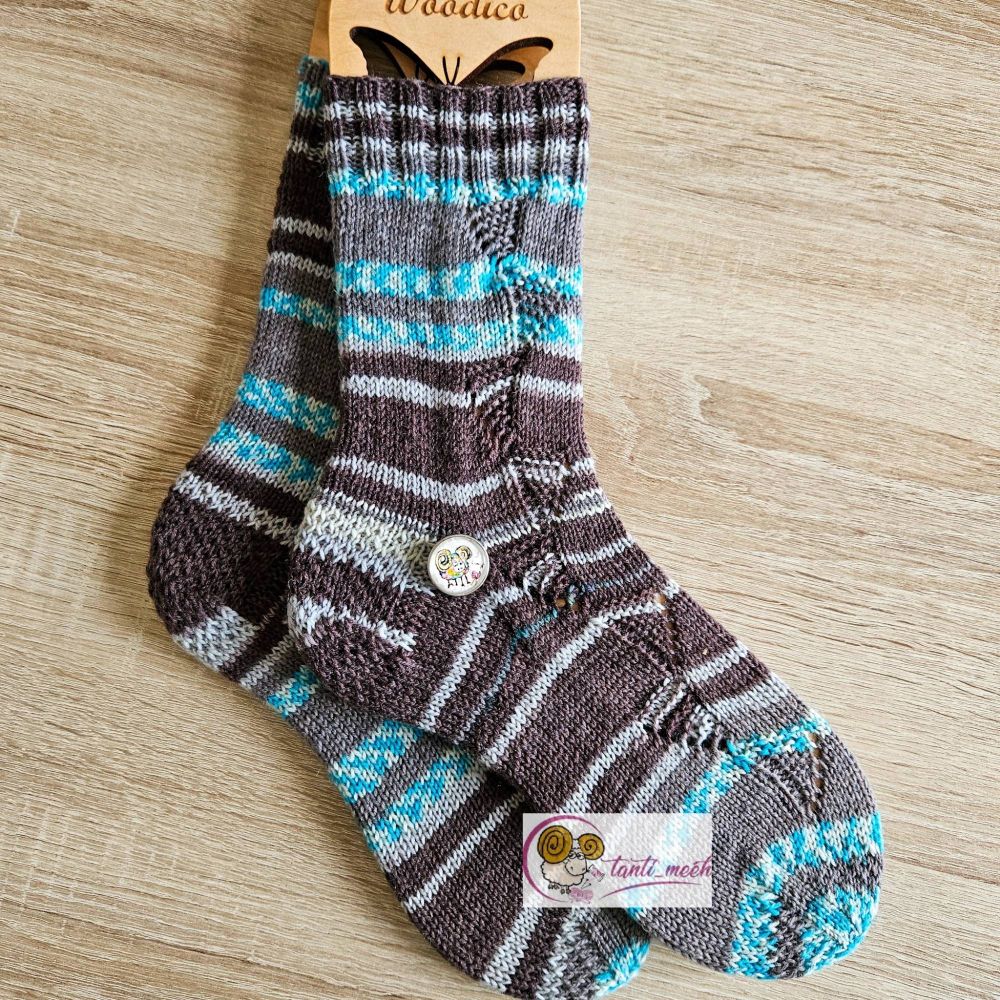 Gestrickte Socken auf Sockenbretter gespannt mit einem Dreickmuster in petrol und grau. Durch den Farbverlauf entstehen verschiedenen grosse Streifen und Blockabschbitte der Farben. Auf Höhe des Fersenanfang liegt ein Druckknopf mit einem bunten, handgemalten Schaf. Die Socken liegen auf einem hellen Tisch.