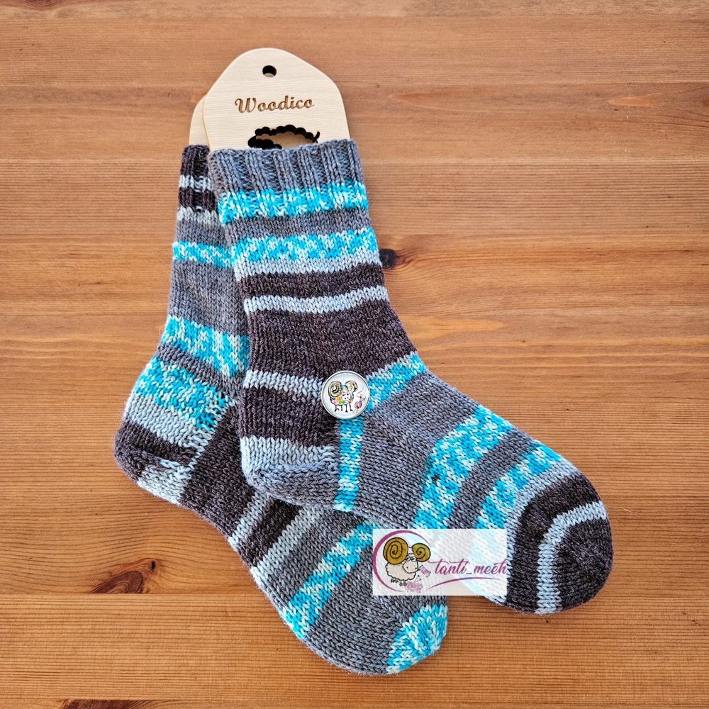 Gestrickte Socken auf Sockenbretter gespannt im Farbverlauf frau ubd petrol. Das Strickbild zeigt verschieden breite Streifen und Blöcke. Auf Höhe des Fersenanfang liegt ein Druckknopf mit einem bunten, handgemalten Schaf.