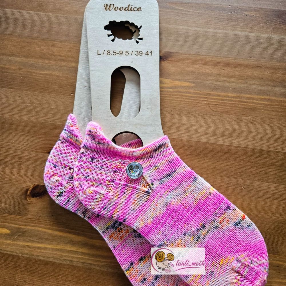 Gestrickte Sneakersocken auf Sockenbretter gespannt in neon pink, mit kleinen Akzenten in gelb. Auf Höhe des Fersenanfang liegt ein Druckknopf mit einem bunten, handgemalten Schaf. Die Sockenbretter liegen auf einem Kiefertisch.