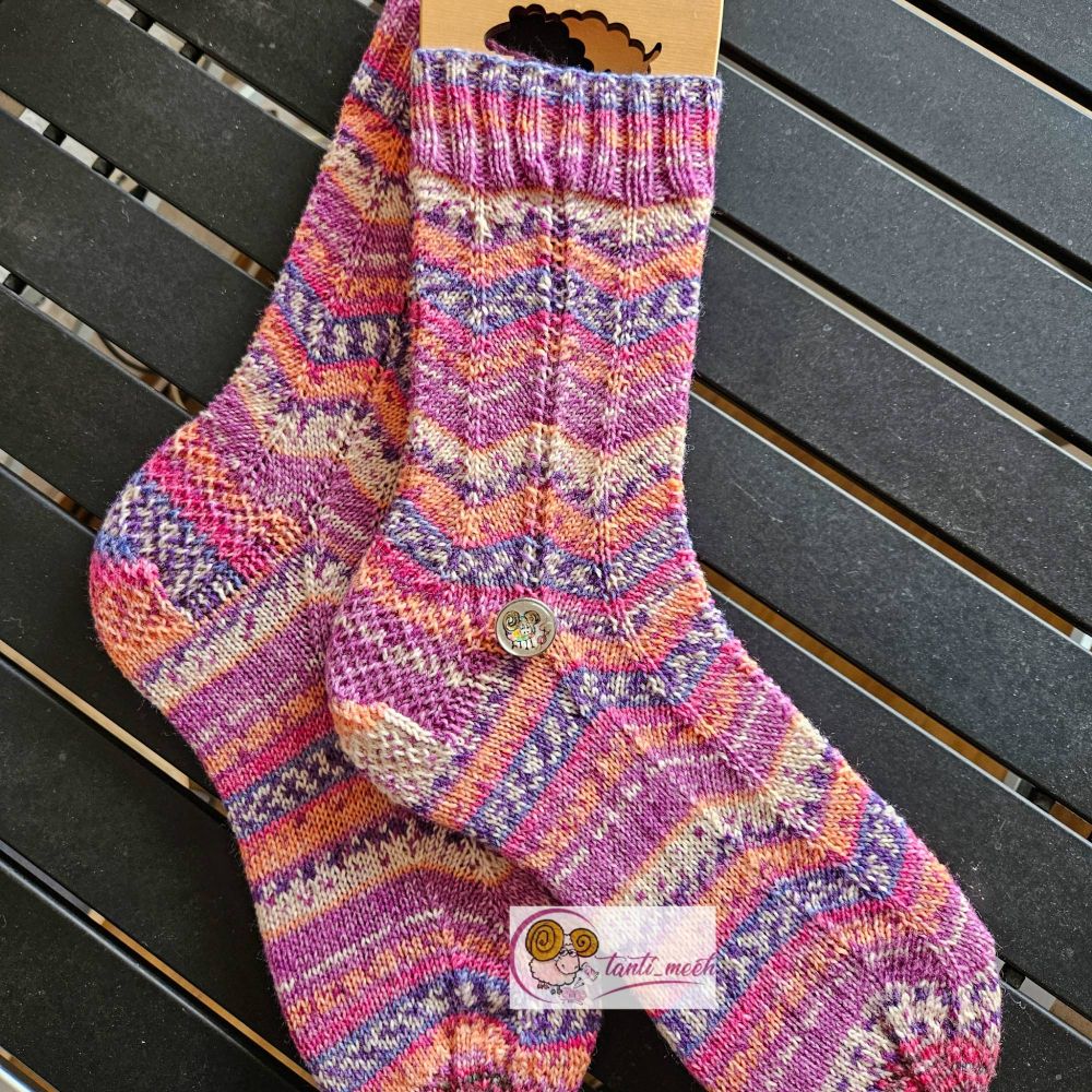 Gestrickte Socken auf Sockenbretter gespannt in verschiedenen Lavendel- ubd Apricottönen. Das Strickbild zeigt ein Zickzackmuster. Auf Höhe des Fersenanfang liegt ein Druckknopf mit einem bunten, handgemalten Schaf. Die Socken liegen auf einem schwarzen Campingtisch.