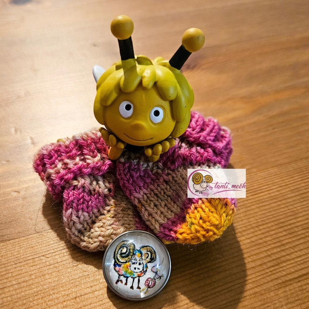 Ganz kleine Söckchen in beige, ros und orange. Das Strickbild zeigt grössere Steifen. Am Bündchen ist eine Biene Maja Figur (Tonies) zu sehen. Unten an der Sockenspitze ist ein Druckknopf mit einem bunten, handgemalten Schaf. Die Söckchen liegen auf einem Kiefertisch.