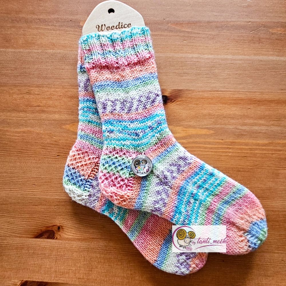 Gestrickte Socken auf Sockenbretter gespannt im Farbverlauf in pastell apricot, grün, gelb. Auf Höhe des Fersenanfang liegt ein Druckknopf mit einem bunten, handgemalten Schaf. Die Socken liegen auf einem Kiefertisch.