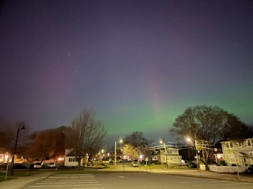 An empty parking lot with a slice of green light just above the horizon, and pale pinkish streaks going upwards above it. 
